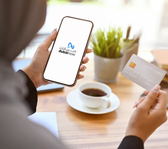Image of a woman sitting at a table, holding a credit card in one hand and her mobile phone in the other. The phone is displaying the Dubai Now app, which is used to make online payments. The woman is drinking a cup of coffee. The image is next to a paragraph about online payment solutions in Dubai.