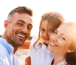 A Western family of three takes a selfie in the summer sun. The parents are smiling and the daughter is laughing, her blonde hair blowing in the wind. The image captures the joy of living in Dubai.